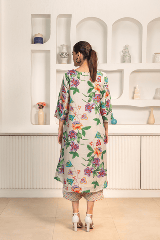 Back view of  woman is wearing a light-colored floral-printed kurta set paired with patterned pants. She is posing against a minimalist white background featuring built-in shelves decorated with vases and small decorative items.