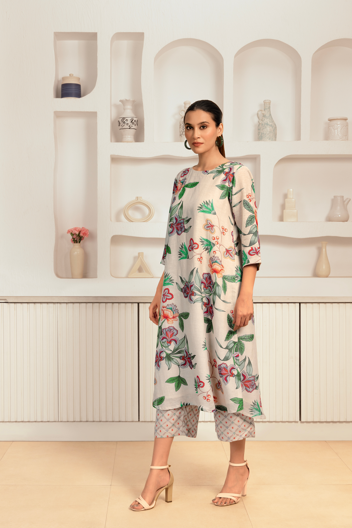 A woman stands gracefully in a floral-patterned kurta and palazzo, surrounded by decorative shelves with various ceramics and flowers.