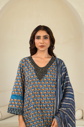 Close up view of a woman wearing a patterned blue and orange traditional outfit with matching pants and a dupatta, standing in front of an arched alcove in a neutral-toned interior.