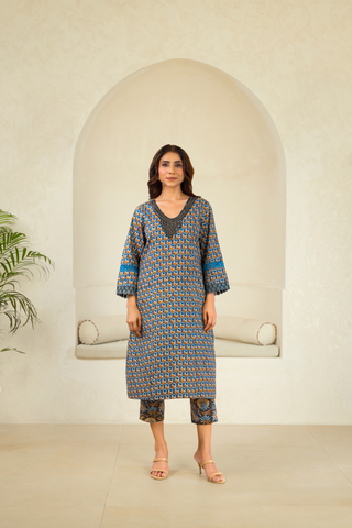 A woman is dressed in a patterned blue and orange traditional outfit with matching pants and a dupatta, standing in front of an arched alcove in a neutral-toned interior.
