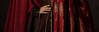 A close-up of a person wearing an ornate dark red and black anarkali kurta set with gold embroidered patterns and a matching draped cloak, shown against a dark background.