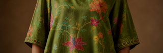 Close-up of a person wearing a loose olive green kurta set with colorful floral embroidery and patterned short sleeves against a warm brown background.