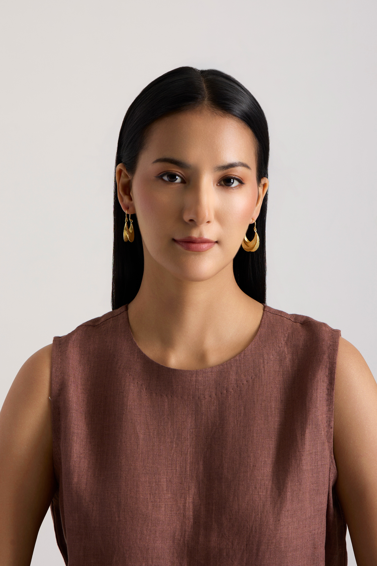 A woman with long dark hair wearing a sleeveless brown top and gold hoop earrings, photographed against a plain light gray background.