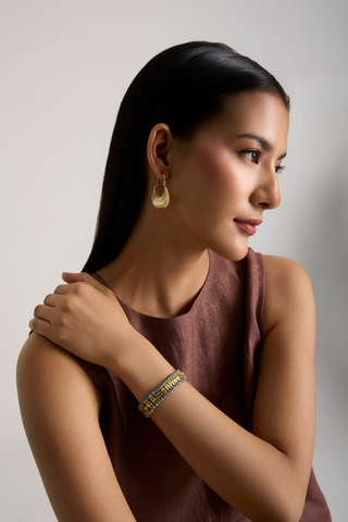 A woman in a sleeveless mauve-brown top wearing a gold crescent-shaped earring and a gold beaded bracelet, shown in profile against a light gray background.