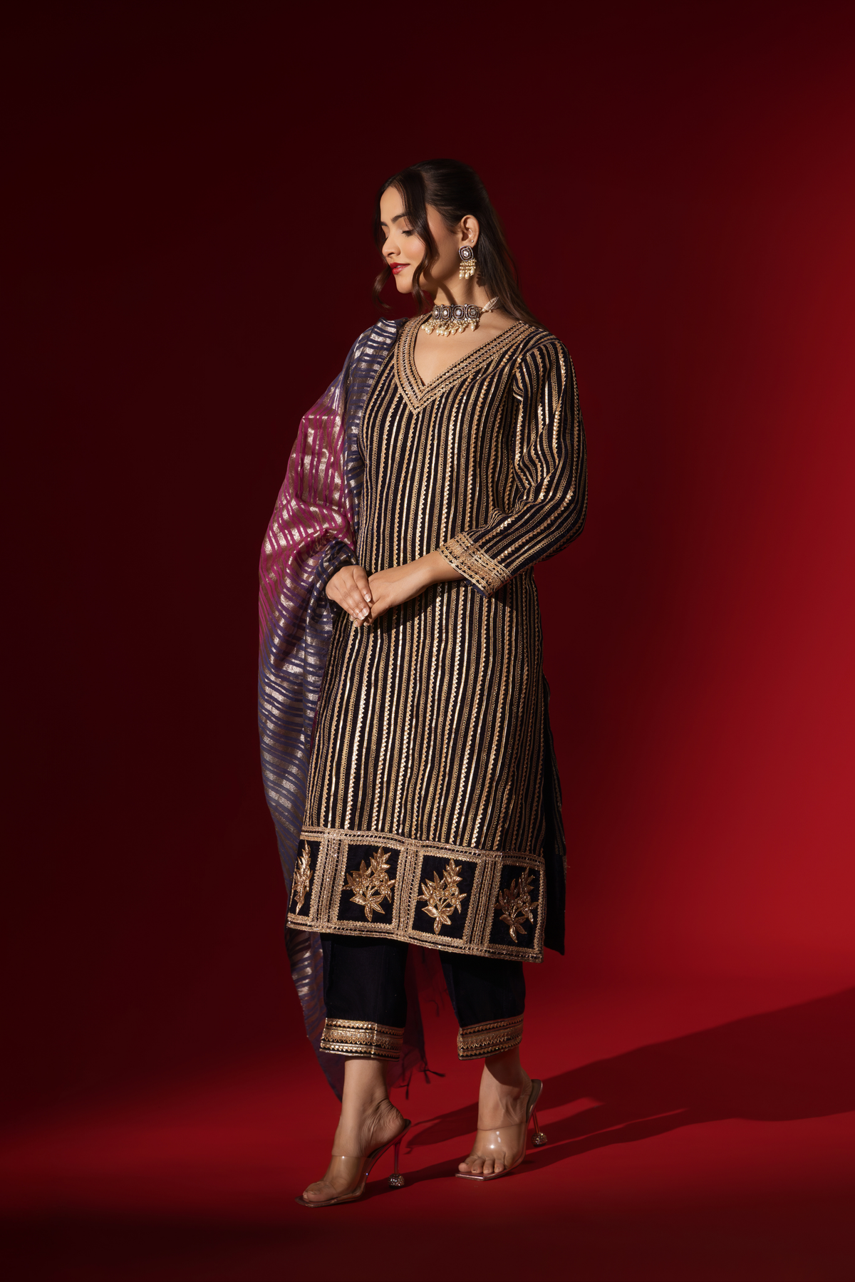 Side view of a woman's hand in an ethnic plum velvet gota kurta set with golden stripes, paired with a shimmering shawl, against a rich red backdrop.