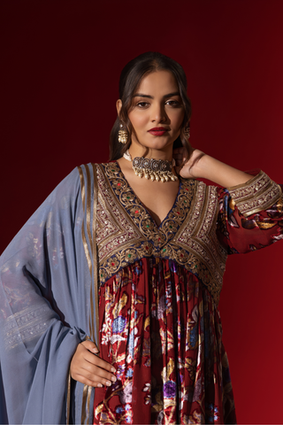 Closeup view of a woman in traditional attire features a richly embellished maroon and floral dress, paired with a blue shawl, against a deep red background.