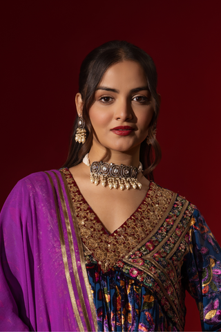Closeup view of a woman stands confidently in a vibrant, embroidered purple printed velvet anarkali kurta set, paired with a matching dupatta against a rich red backdrop.