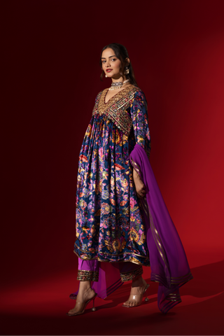 Side view of a woman stands confidently in a vibrant, embroidered purple printed velvet anarkali kurta set, paired with a matching dupatta against a rich red backdrop.