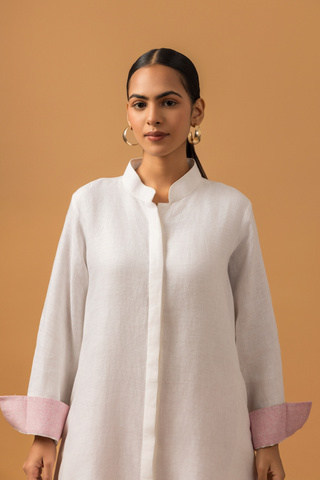 Closeup view of a model wearing a white lotus print linen co-ord set with floral patterns at the hem, paired with pink trousers and standing against a warm orange backdrop.