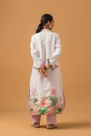 Back view of a model wearing a white lotus print linen co-ord set with floral patterns at the hem, paired with pink trousers and standing against a warm orange backdrop.