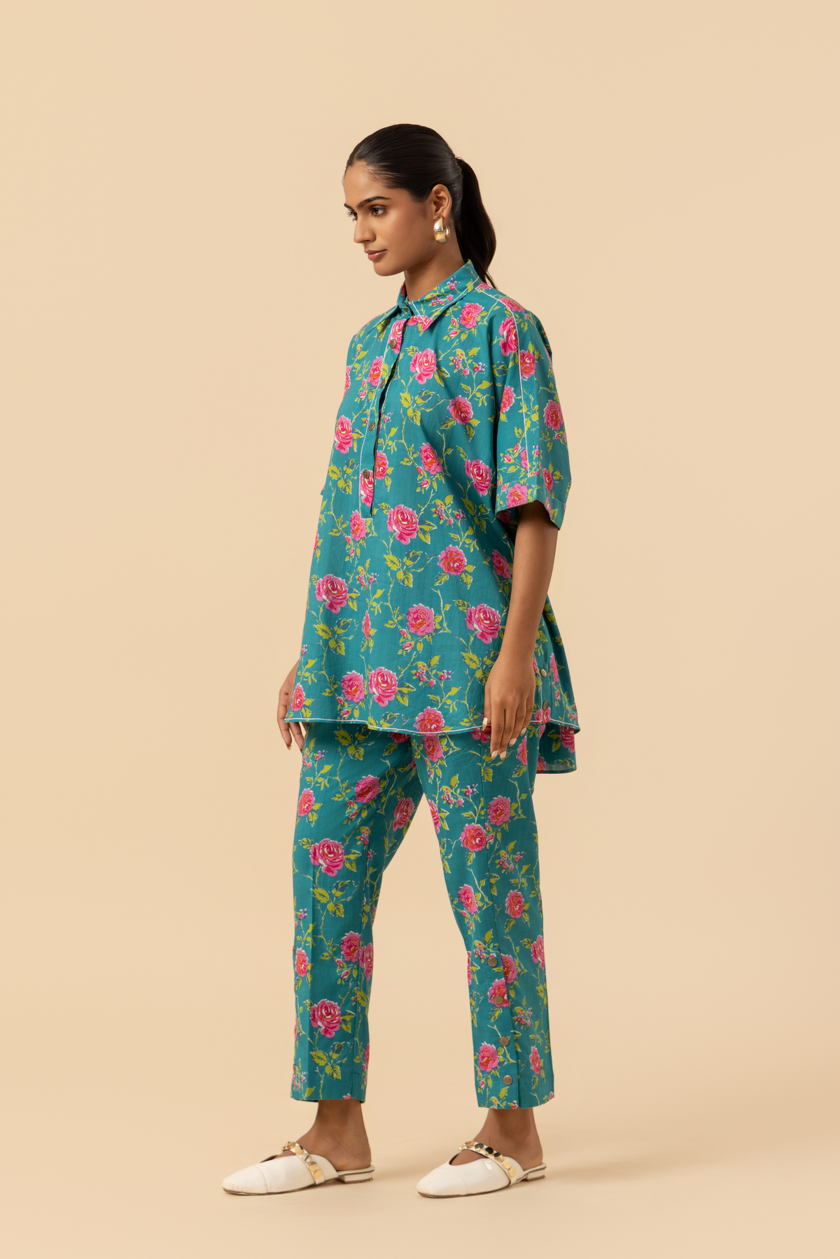 Side vie of a person models a vibrant floral outfit, featuring a teal shirt and matching pants adorned with pink roses, set against a neutral background.