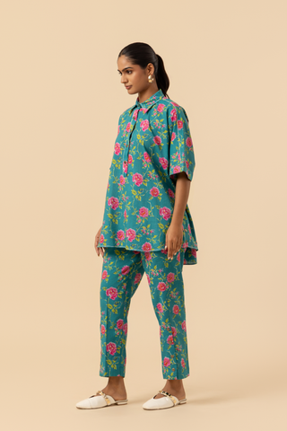 Side vie of a person models a vibrant floral outfit, featuring a teal shirt and matching pants adorned with pink roses, set against a neutral background.