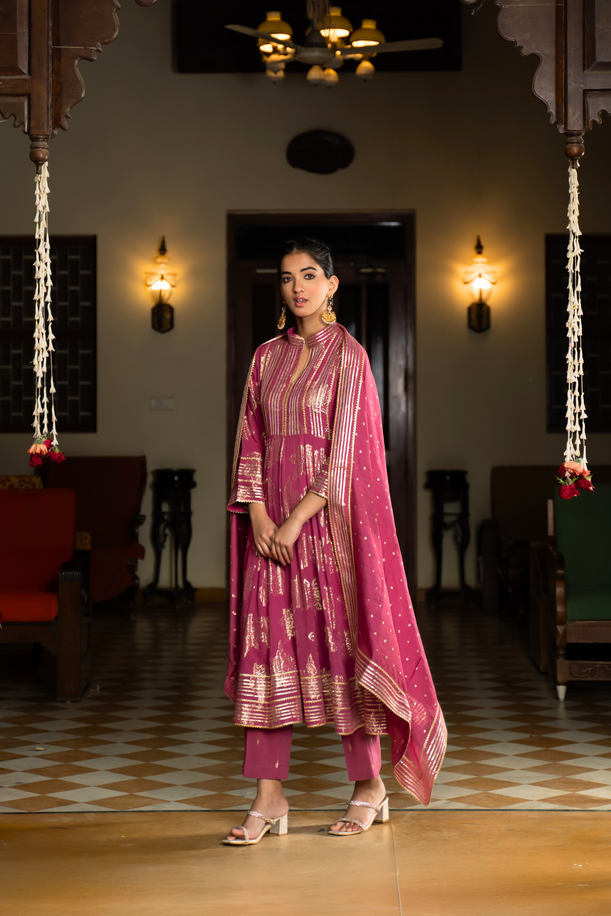 Side view of a  woman in a dark pink traditional anarkali kurta with intricate gold patterns, standing in a beautifully decorated interior.