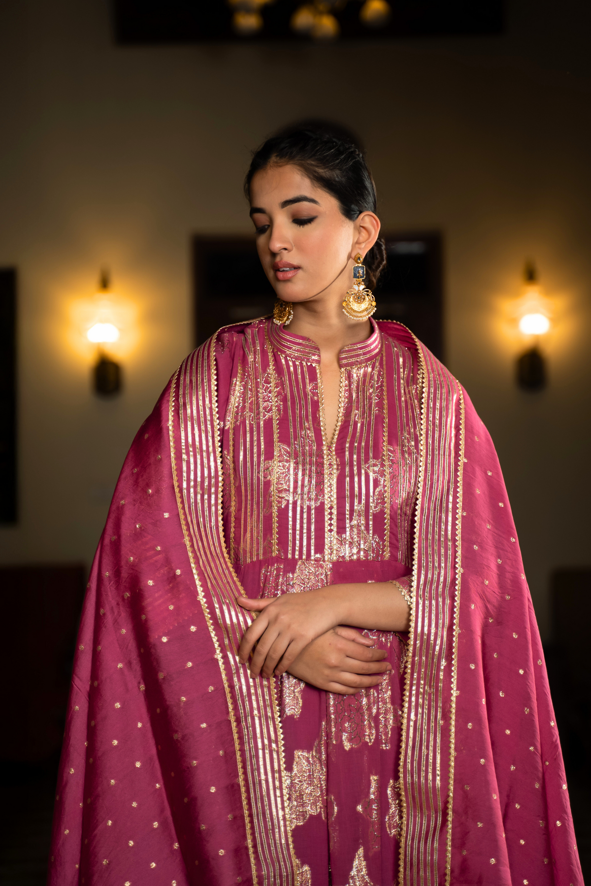 Closeup view of a  woman in a dark pink traditional anarkali kurta with intricate gold patterns, standing in a beautifully decorated interior.
