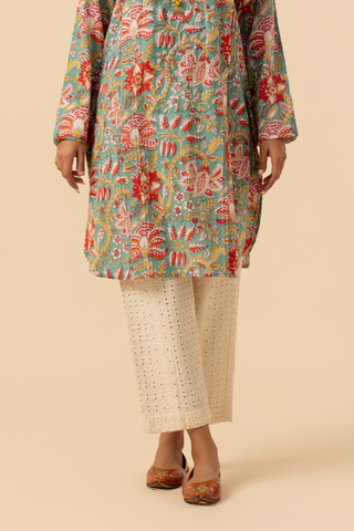A person stands person stands elegantly in a colorful floral co-ord set paired with white embellished pants and traditional sandals against a neutral backdrop.