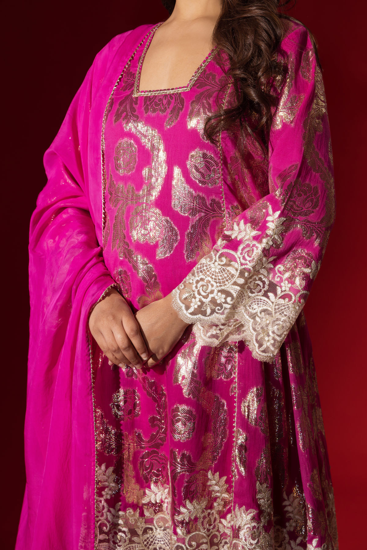 A closeup of woman's hand in an elegant pink shimmer kurta set with intricate golden embroidery stands against a vibrant red background.