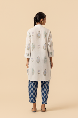 Back view of a woman stands wearing a light-colored, floral-printed kurta paired with blue patterned pants and sandals, against a beige background.