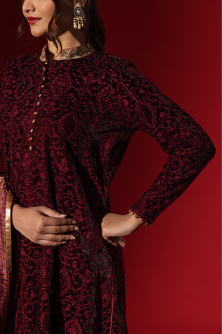 Closeup view of a person's hand in a rich, red traditional outfit adorned with intricate patterns and a shimmering gold-accented shawl against a red backdrop.