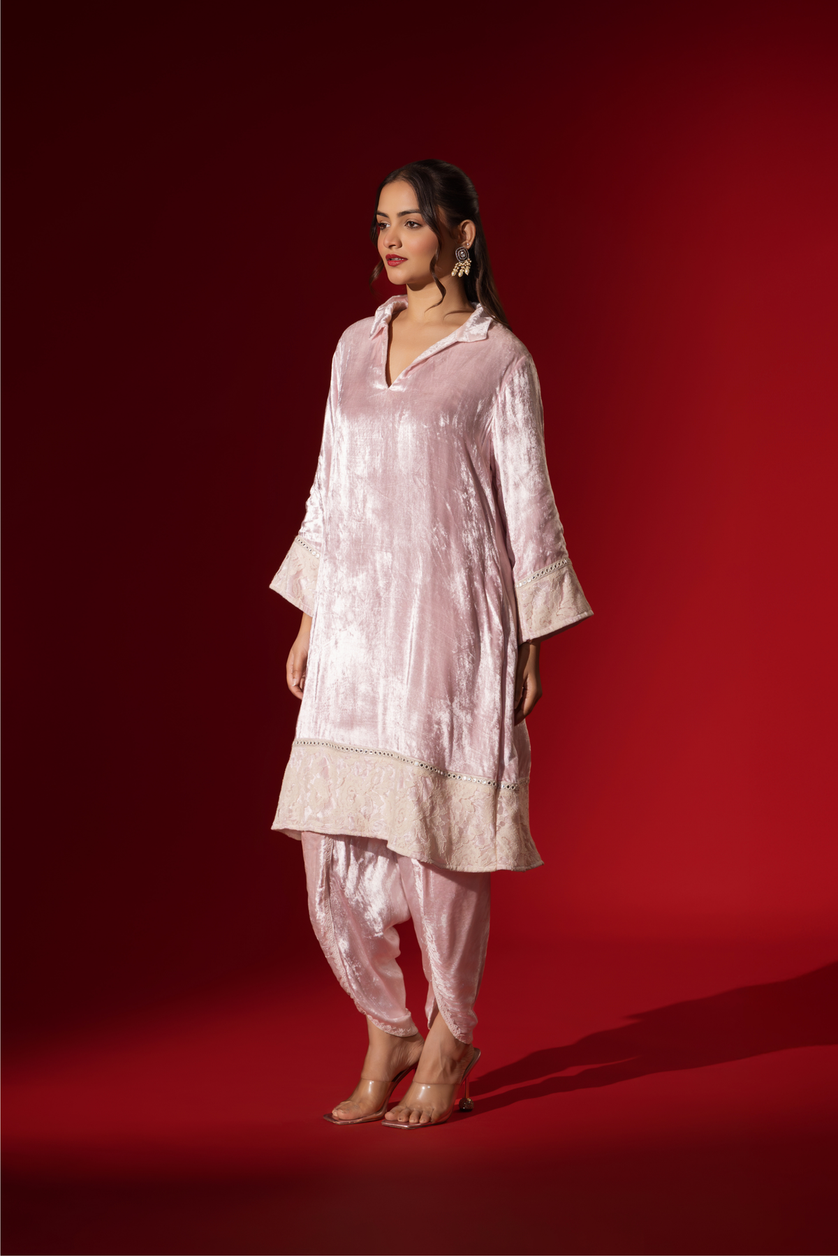 Side view of a woman stands in a champagne pink velvet co-ord set with a long tunic and draped pants, against a vibrant red backdrop.