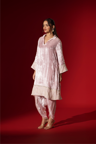 Side view of a woman stands in a champagne pink velvet co-ord set with a long tunic and draped pants, against a vibrant red backdrop.