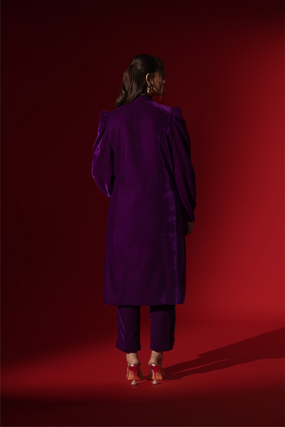 Back view of a woman in a vibrant purple velvet co-ord set with a feminine silhouette and elegant earrings, set against a rich red background.