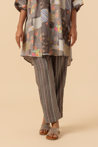 A person stands wearing a floral-patterned oversized tunic paired with striped pants and embellished slide sandals against a neutral backdrop.