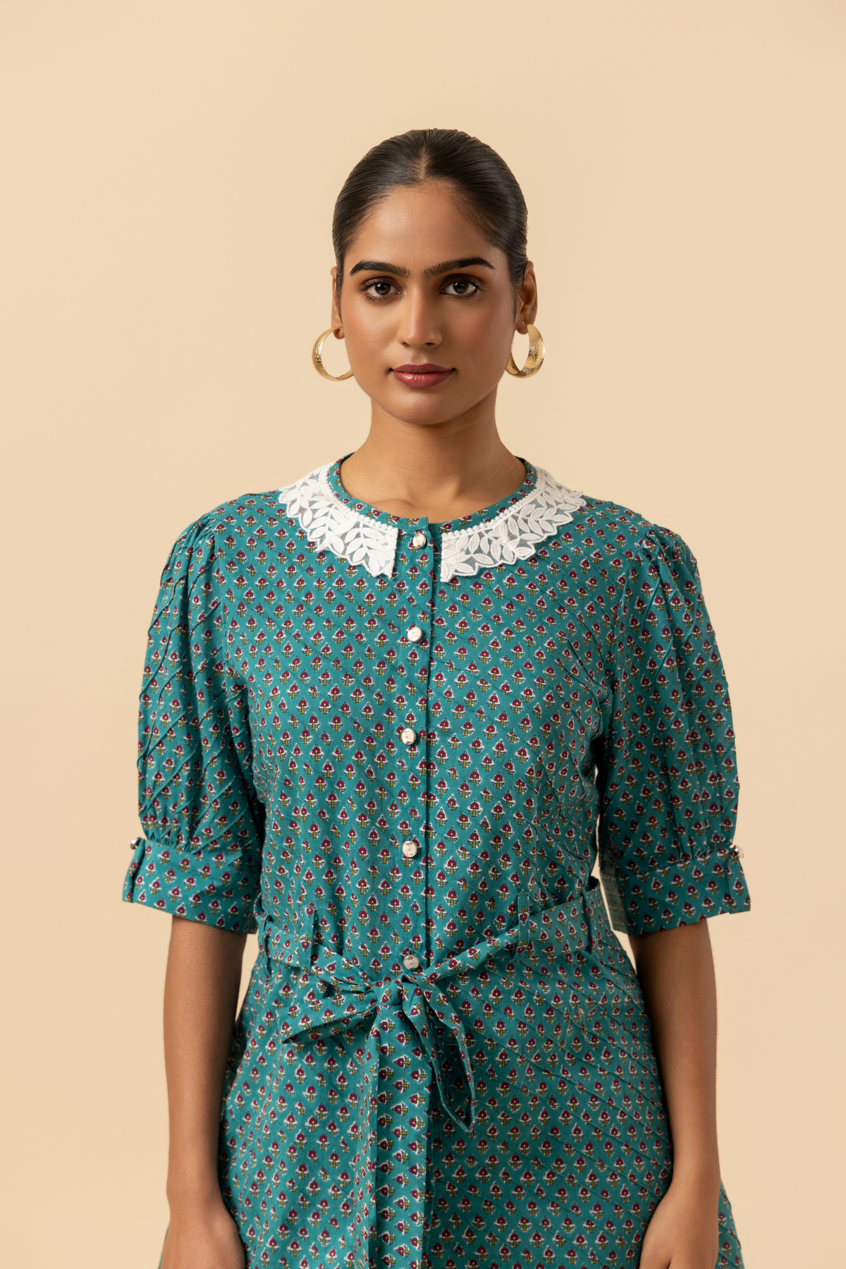 Closeup view of a person stands confidently, wearing a teal patterned outfit with a cinched waist and a collar, set against a neutral background.
