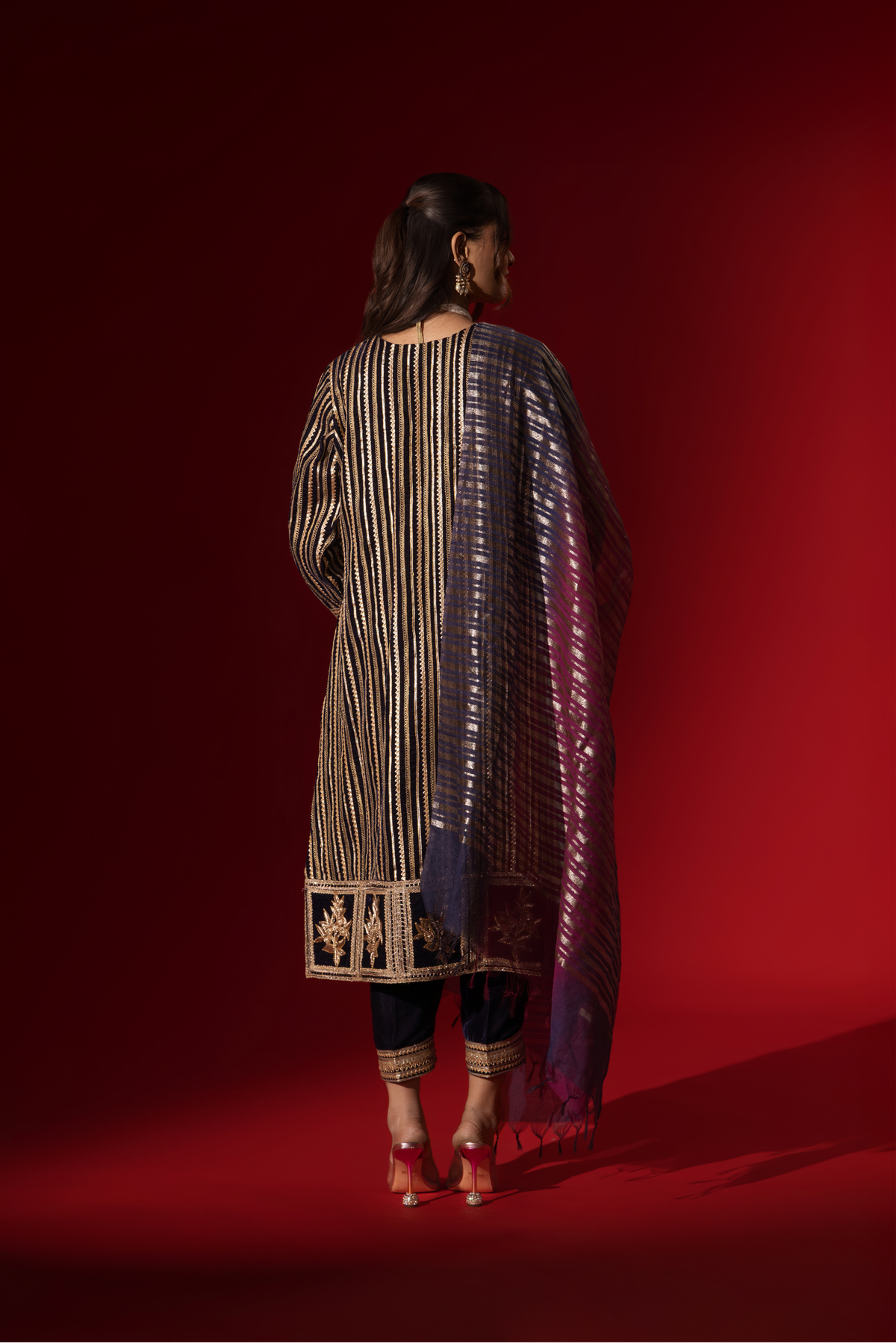 Back view of a woman's hand in an ethnic plum velvet gota kurta set with golden stripes, paired with a shimmering shawl, against a rich red backdrop.
