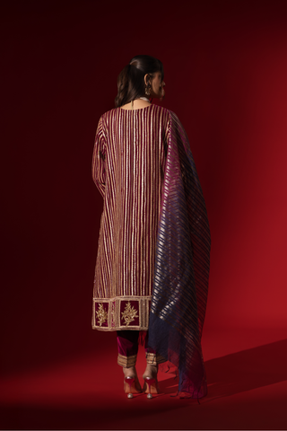 Back view of a woman showcases an ethnic wine velvet gota kurta set with golden stripes, paired with a shimmering shawl, against a rich red backdrop.