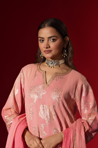 Closeup view of a model in a flowing pink ethnic outfit adorned with intricate golden patterns, standing against a deep red backdrop.