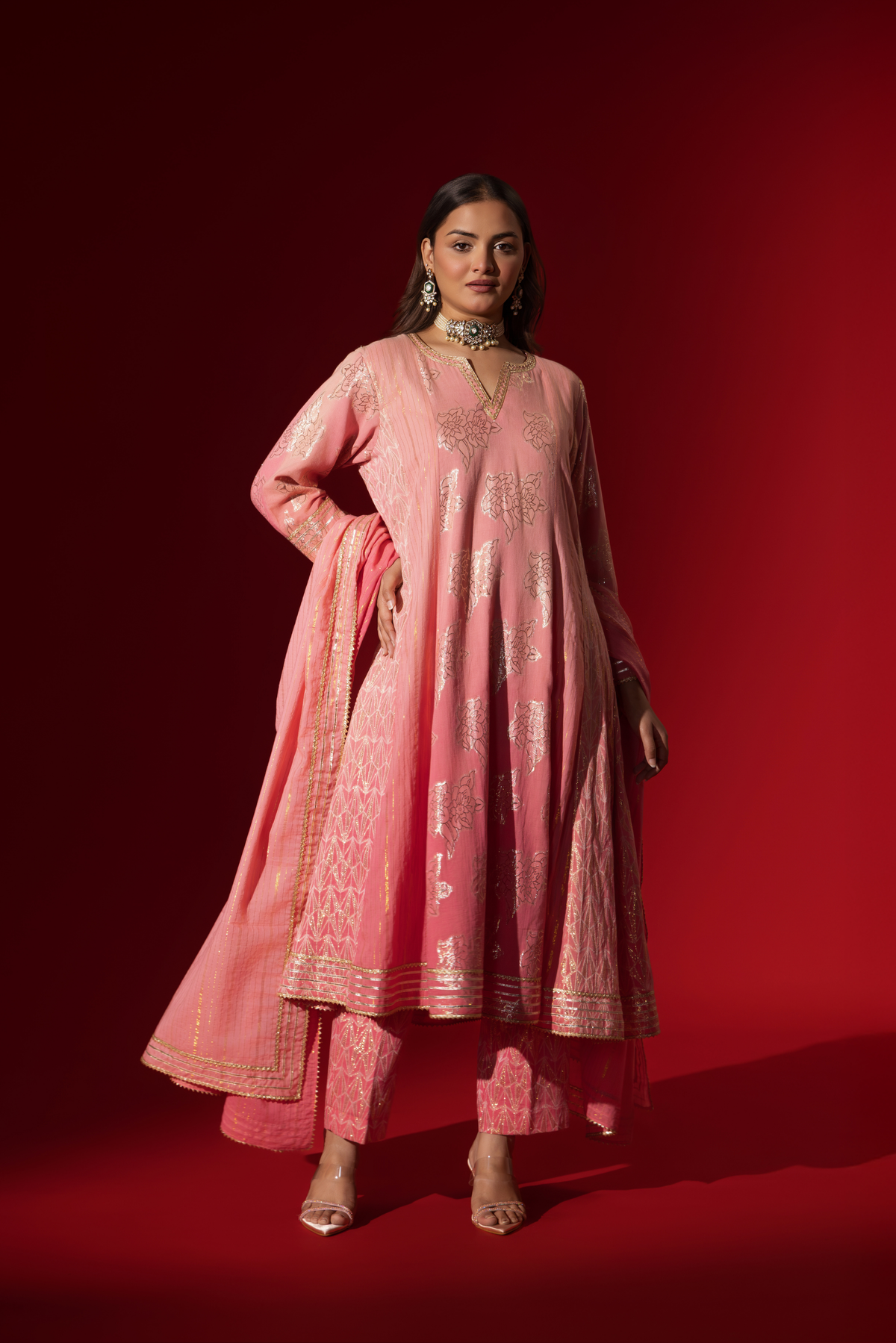 A model in a flowing pink ethnic outfit adorned with intricate golden patterns, standing against a deep red backdrop.