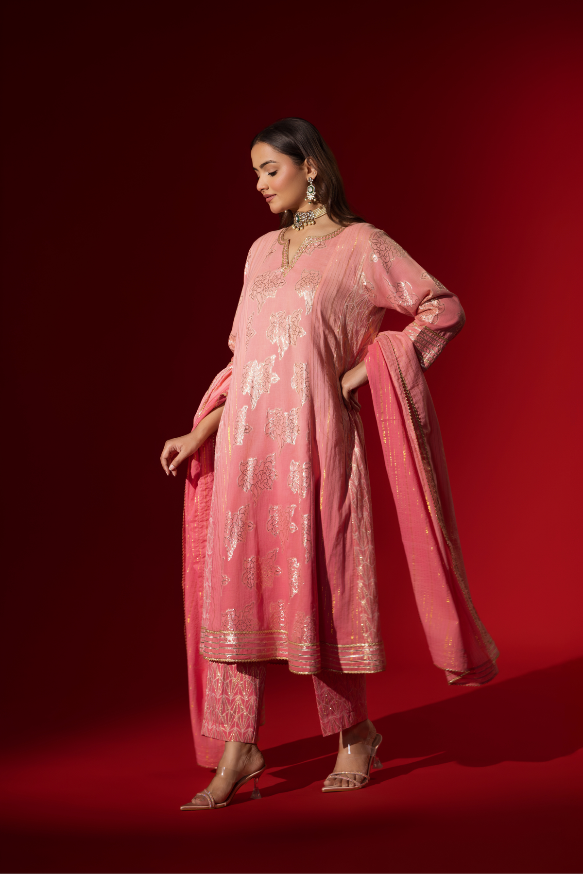 Side view of a model in a flowing pink ethnic outfit adorned with intricate golden patterns, standing against a deep red backdrop.