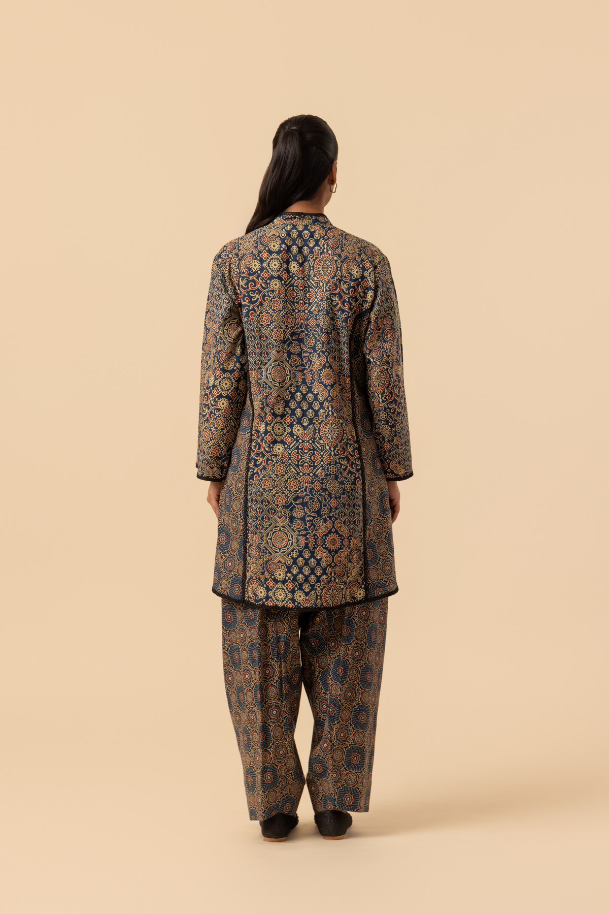 Back view of a model wearing a long, patterned ajrakh print co-ord set which has tunic and loose pants, showcasing intricate designs in dark blues and golds against a neutral background.