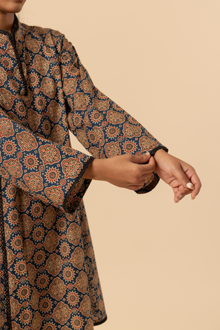 Closeup view of a woman's hand wearing a patterned ajrakh print farshi co-ord set which has tunic with matching trousers, showcasing vibrant colors and intricate designs against a simple beige background.