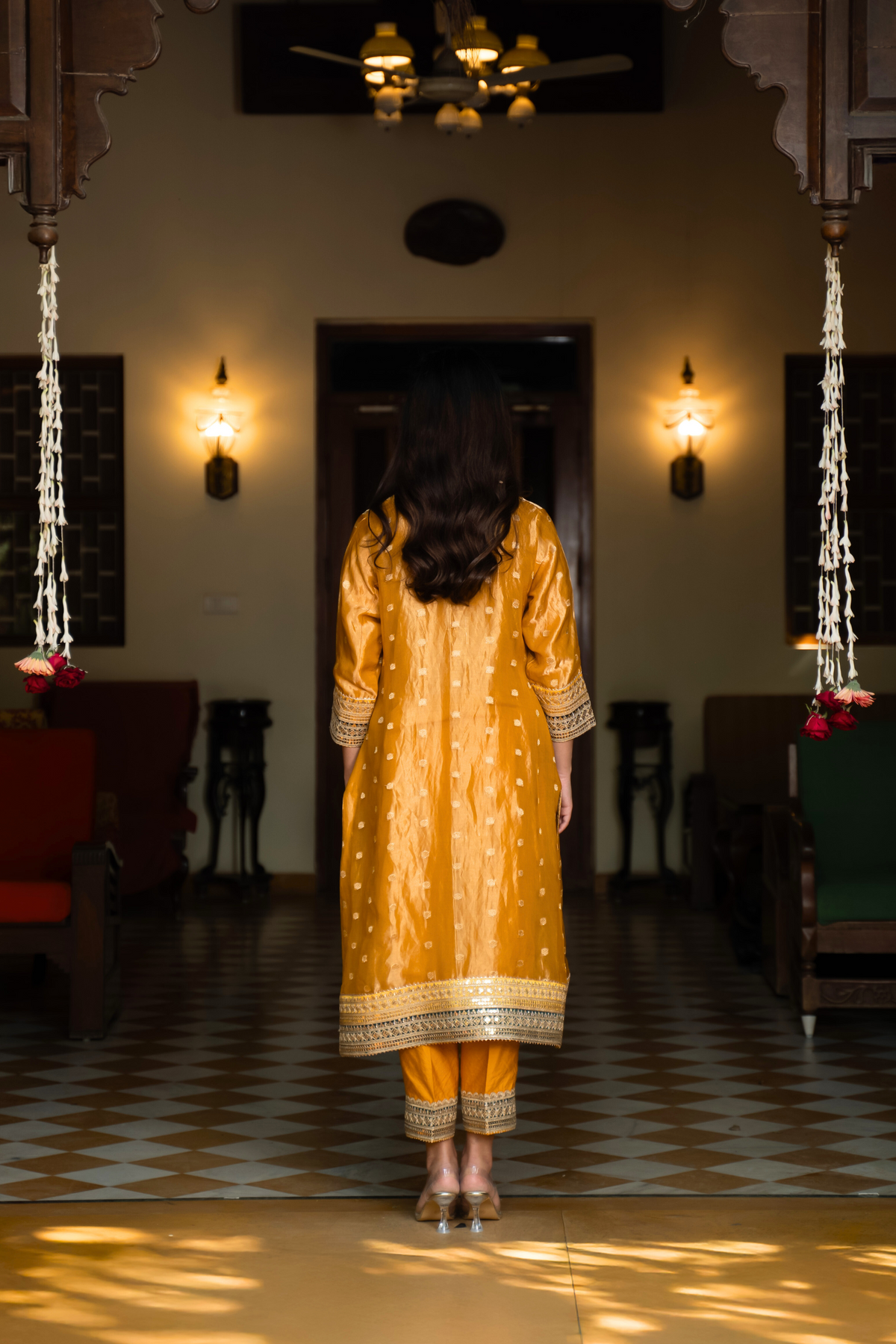 Back view of a  person wearing a vibrant yellow traditional banarsi silk kurta set stands in an elegant room with vintage decor and warm lighting.