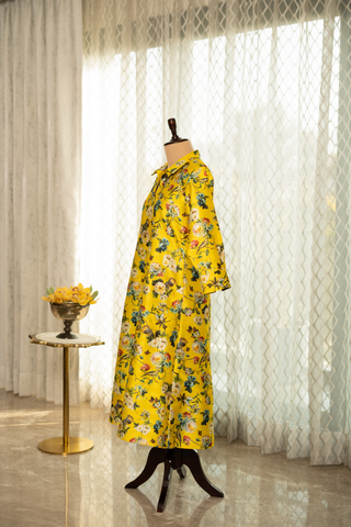 Side View of a  vibrant yellow floral dress displayed on a mannequin stand, set in a room with soft natural lighting, white curtains, and a decorative side table with yellow flowers.