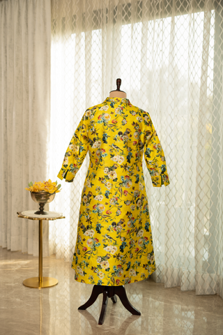Back View of a  vibrant yellow floral dress displayed on a mannequin stand, set in a room with soft natural lighting, white curtains, and a decorative side table with yellow flowers.