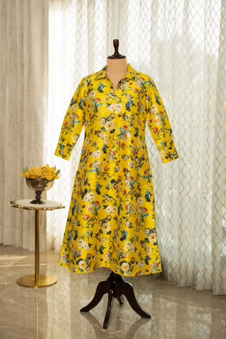 A vibrant yellow floral dress with a collared neckline and three-quarter sleeves displayed on a mannequin in a softly lit room.