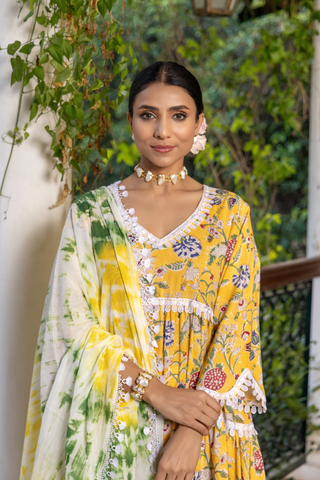 Closeup view of a woman stands gracefully in a vibrant yellow floral cotton kurta set, paired with a light green and white dupatta against a lush, green backdrop.