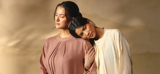 Two women in draped, flowing garments pose closely against a warm beige background. One wears a dusty mauve-brown top, and the other wears an off-white top.