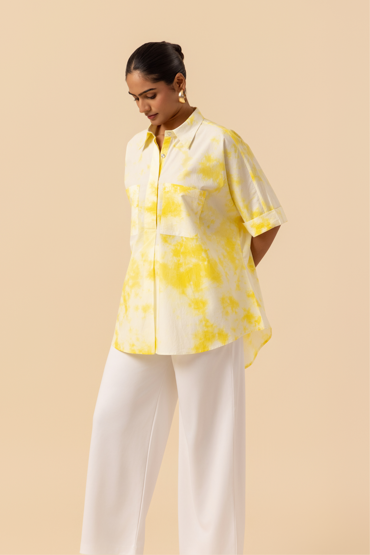 Side view of a model poses confidently in a yellow tie-dye oversized shirt and white wide-leg pants against a soft beige background.
