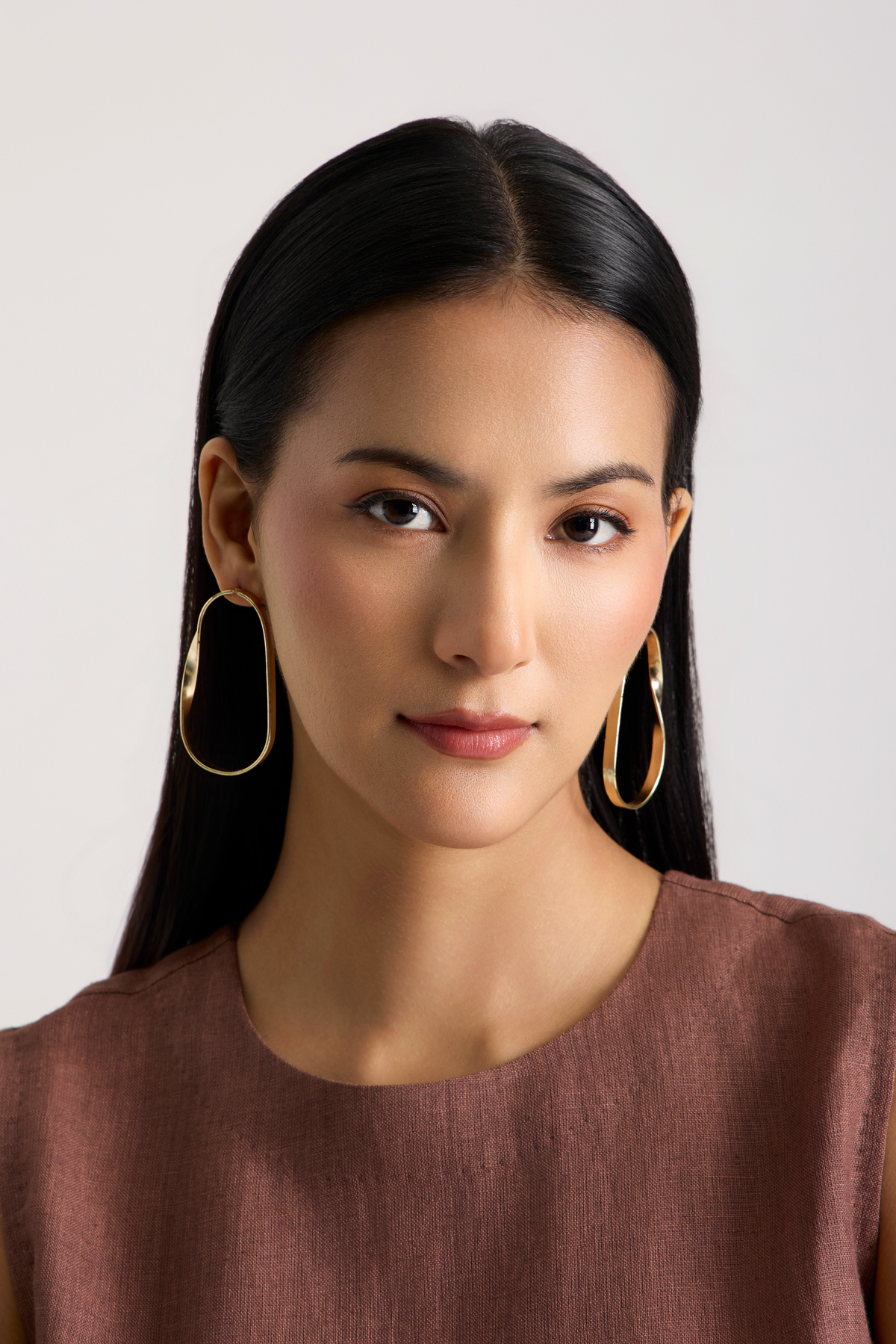 A woman with long black hair wearing large gold-tone oval hoop earrings and a brown top, shown against a plain light gray background.