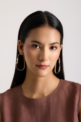 A woman with long black hair wearing large gold-tone oval hoop earrings and a brown top, shown against a plain light gray background.