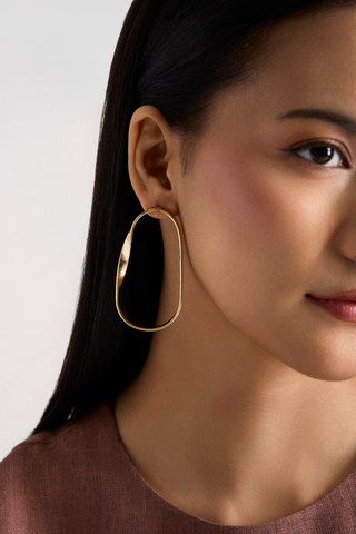 A close-up of a woman wearing a large, polished gold-toned oval hoop earring, shown against a clean light gray background.