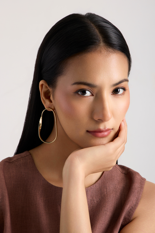 A woman with long black hair posing in a brown sleeveless top, wearing a large gold-toned oval hoop earring against a light gray background.