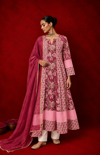 A woman wearing a pink floral printed anarkali kurta with white pants and a matching dupatta, styled with traditional jewelry, posing against a rich red backdrop.