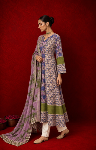 Left side of a woman wearing a purple and green floral printed kurta with white pants and a matching printed dupatta, accessorized with traditional jewelry, posing against a rich red backdrop.